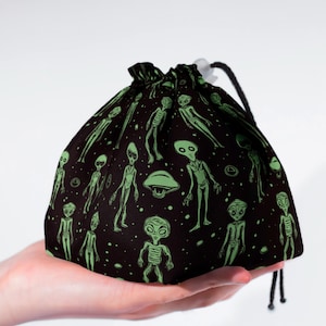 Può includere: Borsa nera con coulisse, decorata con un motivo ripetuto di figure aliene verdi e dischi volanti. La borsa è tenuta in mano e ha una chiusura con coulisse nera. Gli alieni sono in varie pose.