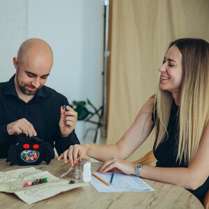 Puede incluir: Una bolsa de dados negra con forma de ara&ntilde;a con ojos rojos, llena de dados coloridos. Un hombre y una mujer est&aacute;n sentados a una mesa, con un mapa y &uacute;tiles de dibujo. La mujer sonr&iacute;e.