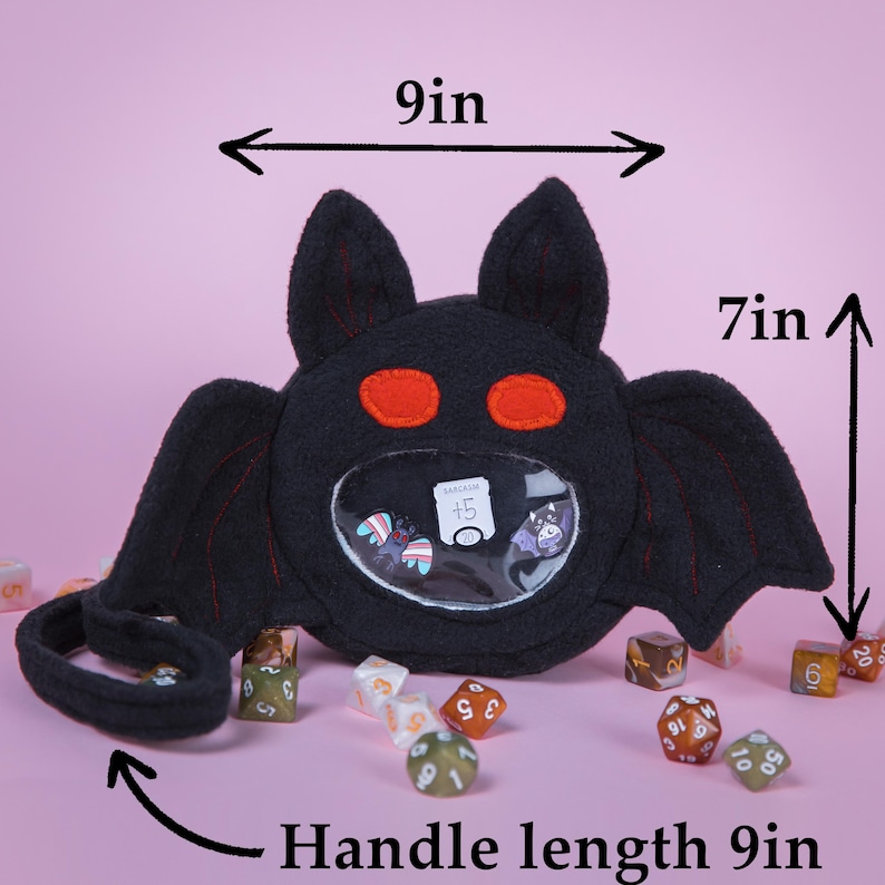 Puede incluir: Una bolsa negra con forma de murci&eacute;lago con ojos rojos y un bolsillo frontal transparente. La bolsa mide 23 cm de ancho, 18 cm de alto y tiene un asa de 23 cm. El bolsillo contiene dados y una tarjeta "Sarcasm +5".