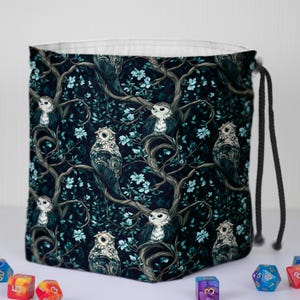 May include: A square, drawstring bag with a repeating pattern of owls perched on tree branches with blue flowers. The background is a dark teal color. The bag has a white interior and a black drawstring.