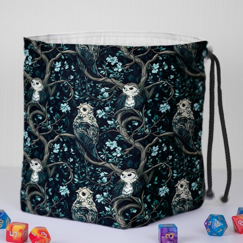 May include: A square, drawstring bag with a repeating pattern of owls perched on tree branches with blue flowers. The background is a dark teal color. The bag has a white interior and a black drawstring.