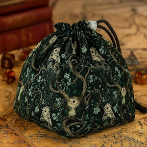 May include: A drawstring dice bag with a dark green background and a repeating pattern of owls, trees, and foliage. The bag is made of fabric and has a black drawstring closure. The bag is sitting on a map.