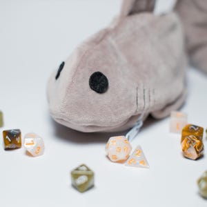 May include: A plush shark-shaped dice bag in a soft gray color, with black button eyes and stitched details. Scattered around the bag are various polyhedral dice in shades of white, brown, and gold, perfect for tabletop gaming.