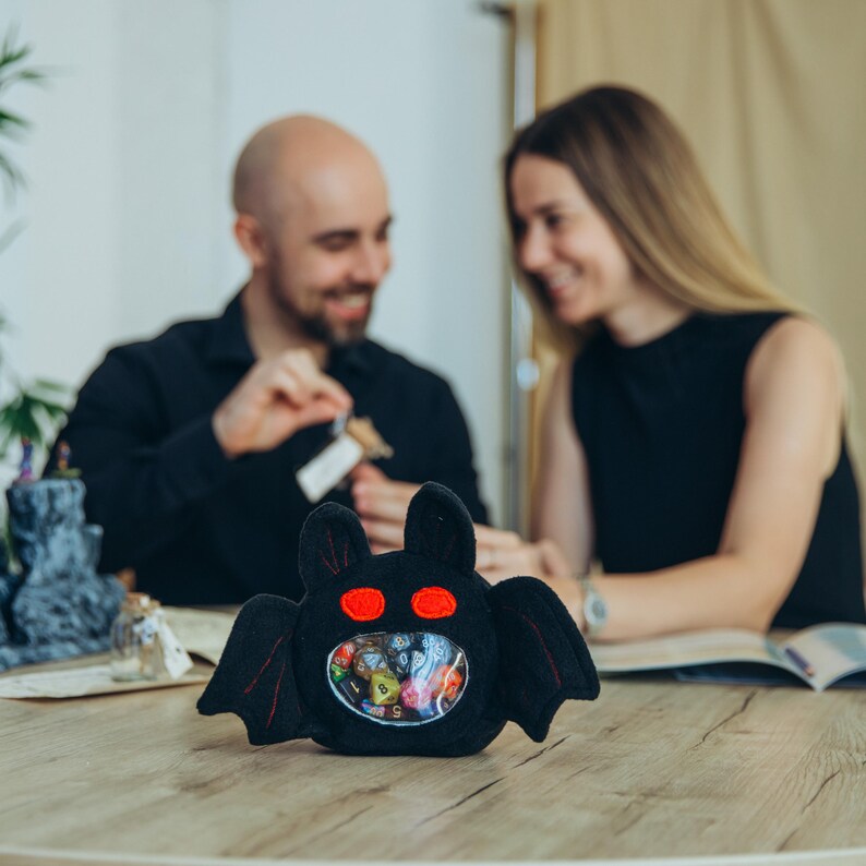 Puede incluir: Una bolsa de dados de felpa negra con forma de murci&eacute;lago con ojos rojos y una barriga transparente, llena de dados de colores. El murci&eacute;lago tiene alas y est&aacute; sobre una mesa de madera. Una pareja est&aacute; en el fondo.