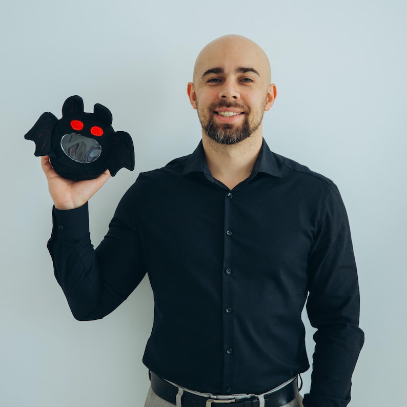 Puede incluir: Una bolsa negra de felpa con forma de murci&eacute;lago con ojos rojos y un centro claro y circular. La bolsa tiene una cara sonriente y se sostiene sobre un fondo blanco liso. Accesorio tem&aacute;tico de Halloween.