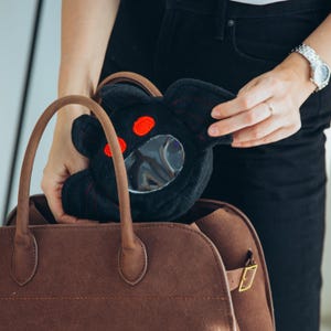 Puede incluir: Una bolsa de felpa negra con forma de personaje de dibujos animados, con ojos rojos y una ventana circular transparente. Se est&aacute; colocando en un bolso tote marr&oacute;n con asa.