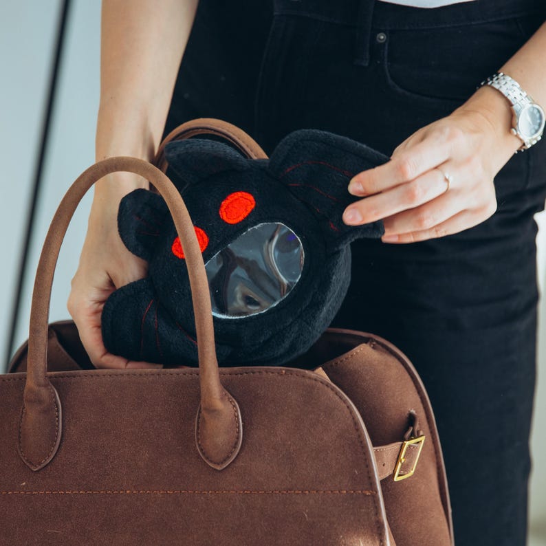 Puede incluir: Una bolsa de felpa negra con forma de personaje de dibujos animados, con ojos rojos y una ventana circular transparente. Se est&aacute; colocando en un bolso tote marr&oacute;n con asa.