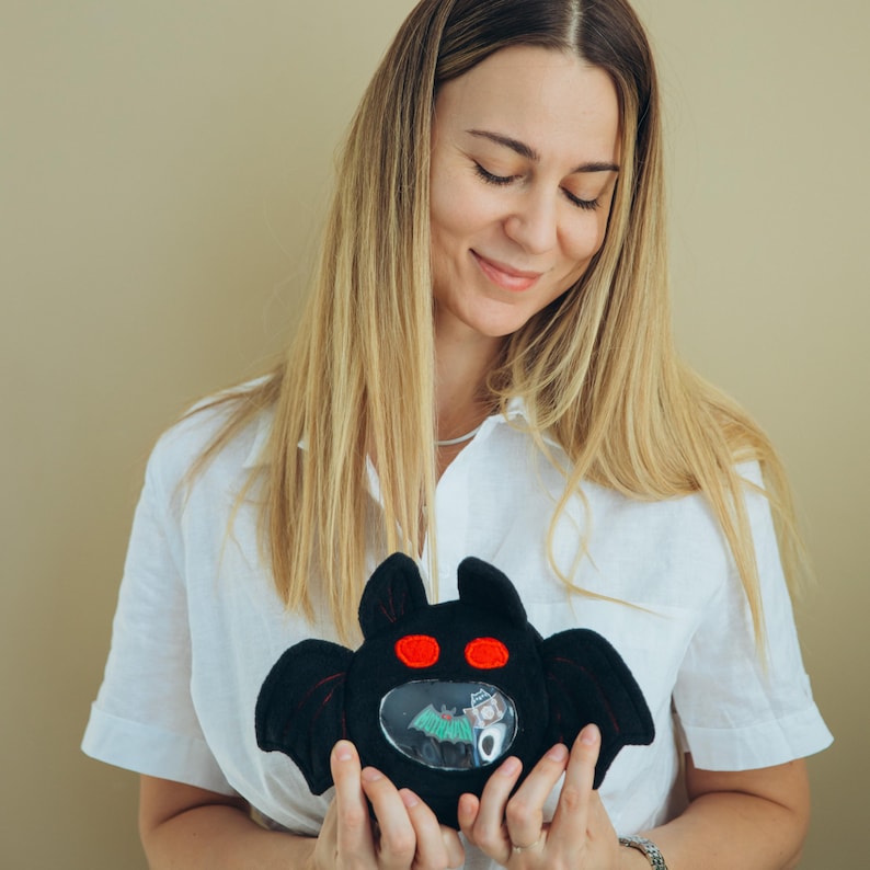 Puede incluir: Un juguete de peluche negro con forma de murci&eacute;lago con ojos rojos y una ventana transparente que muestra una escena de Halloween. El juguete tiene alas extendidas y lo sostiene una persona que lleva una camisa blanca.