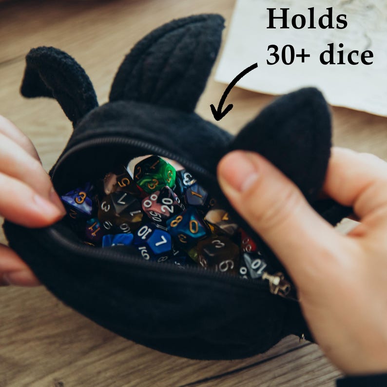 Puede incluir: Bolsa de dados de felpa negra con forma de conejito, con el texto "Holds 30+ dice". La bolsa est&aacute; abierta, revelando una variedad de dados poli&eacute;dricos coloridos. La bolsa tiene dos orejas verticales y un cierre de cremallera.