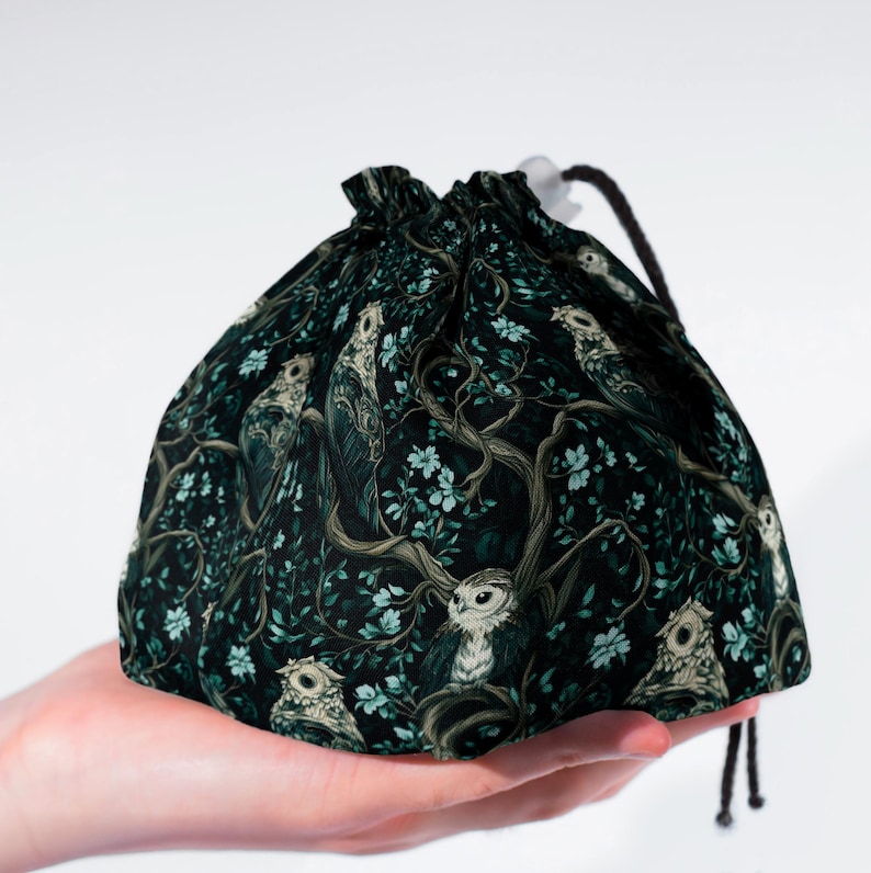 May include: A dark green drawstring bag featuring a repeating pattern of owls, branches, and leaves. The bag has a black drawstring closure and is held in a hand. The design has a whimsical, forest-like theme.