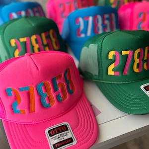 Könnte beinhalten: Ein rosafarbener Trucker-Hut mit einer großen, mehrfarbigen Nummer "27858", die auf der Vorderseite gestickt ist. Der Hut hat ein Netzrücken und ein schwarz-weißes Etikett mit der Aufschrift "OTTO WORLD FAMOUS FOAM FRONT OSFM TRUCKER".