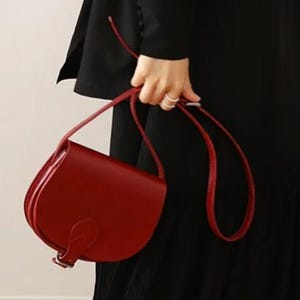 May include: A red leather saddle bag with a buckle closure and a long adjustable strap.