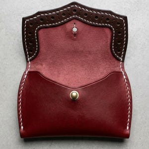 May include: A handcrafted, open leather wallet in a rich burgundy color. The wallet features a dark brown, perforated top edge with white stitching. The interior is burgundy, and the wallet is secured with a gold-colored snap closure.