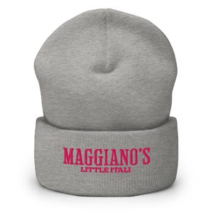 May include: A gray beanie with the embroidered text "Maggiano's Little Italy" in pink. The beanie is a folded, knit cap, suitable for cold weather. The hat is made of a soft, comfortable material.