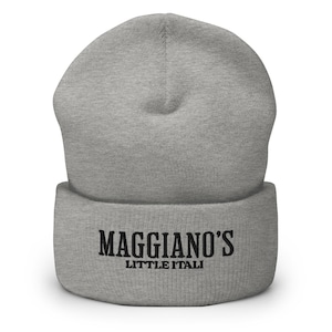 May include: A light gray beanie with the text "Maggiano's Little Italy" in black. The beanie is made of a soft, knitted material and has a folded brim. The text is centered on the brim.