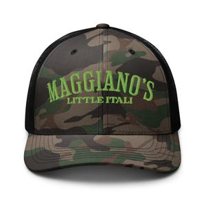 May include: Camouflage trucker hat with the text "Maggiano's Little Italy" in green. The hat features a black mesh back and a curved brim. The front panel has a camouflage pattern in shades of green, brown, and tan.