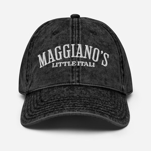 May include: A black, distressed-look baseball cap with the embroidered text "Maggiano's Little Italy" in white. The cap has a curved brim and a button on top. The hat is a classic, casual accessory.