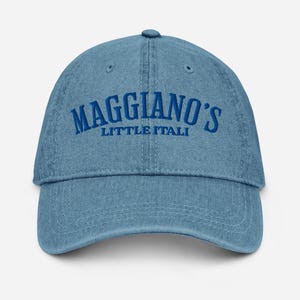 May include: Light blue denim baseball cap with the text "Maggiano's Little Italy" embroidered in blue. The cap has a curved brim and an adjustable strap at the back.