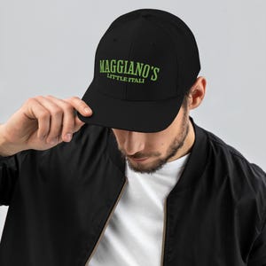 May include: Black baseball cap with a mesh back and the text "Maggiano's Little Italy" in green. The cap is being held by a person wearing a black jacket.