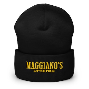 May include: Black beanie with the embroidered text "MAGGIANO'S LITTLE ITALIA" in gold. The beanie is a classic, warm winter hat, perfect for cold weather. The hat is made of a soft, knitted material.