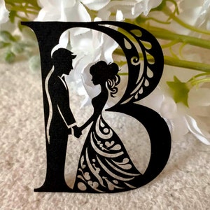 Puede incluir: Corte de papel negro de una novia y un novio de pie uno al lado del otro, tomándose de la mano. La pareja está posicionada frente a la letra "B" que también está cortada de papel negro.
