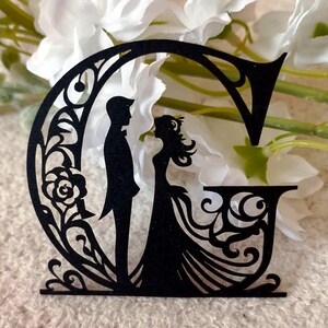 May include: Black paper cut-out of the letter G with a silhouette of a bride and groom inside. The design features intricate floral details.
