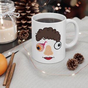 May include: White ceramic coffee mug with a cartoon face printed on it. The face has a brown curly hair, one eye is a cartoon eyeball, the other eye is a bloody eyeball, and a red nose and lips.