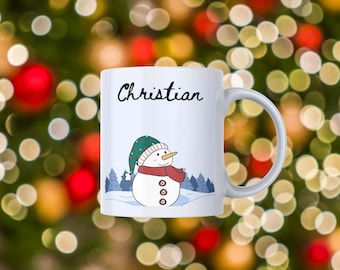 May include: White ceramic mug with the name "Christian" in black script. The mug features a snowman illustration wearing a green hat and red scarf. The background is a blurred image of Christmas lights.