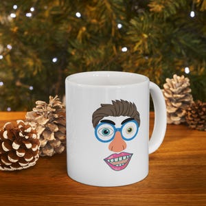 May include: White ceramic mug with a cartoon face printed on it. The face has brown hair, blue glasses, a pink nose, and a pink mouth with white teeth.