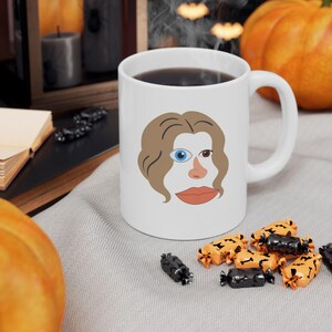 May include: White ceramic coffee mug with a cartoon illustration of a woman's face with one blue eye and one brown eye. The mug is filled with coffee and has a brown handle.
