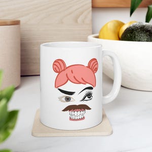 May include: White ceramic mug with a pink cartoon face with one eye open and one eye closed, a mustache, and teeth. The mug has a handle on the right side.