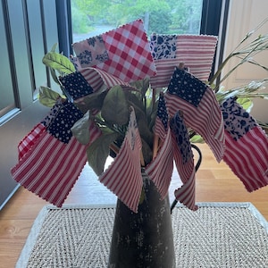 May include: A collection of small fabric flags featuring various red, white, and blue patterns, including gingham, stripes, and floral prints, arranged in a dark metal pitcher. The flags are mounted on wooden sticks and mixed with green foliage.