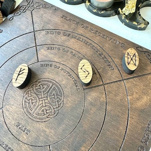K&ouml;nnte beinhalten: Ein dunkles Holz-Divinationsbrett mit eingravierten konzentrischen Kreisen und Text, darunter "PLACE OF RECEIVING" und "RING OF MANIFESTATION". Drei Runensteine aus Holz sind auf dem Brett platziert. Ein schwarz-goldener mondf&ouml;rmiger Kerzenhalter mit Kristallen und einer brennenden Kerze befindet sich im Hintergrund.