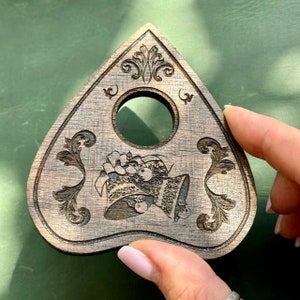 May include: A wooden planchette with a decorative design. The planchette is triangular with a circular viewing hole and ornate floral and bell engravings. The wood has a warm, brown tone. The planchette is held by a person.