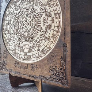 Może przedstawiać: Drewniany, okrągły kalendarz z symbolami astrologicznymi i tarota, w tym znakami zodiaku i nazwami kart. Kalendarz jest grawerowany ze skomplikowanymi detalami i słowami "Blessed Be". Jest wyświetlany na drewnianym stojaku.