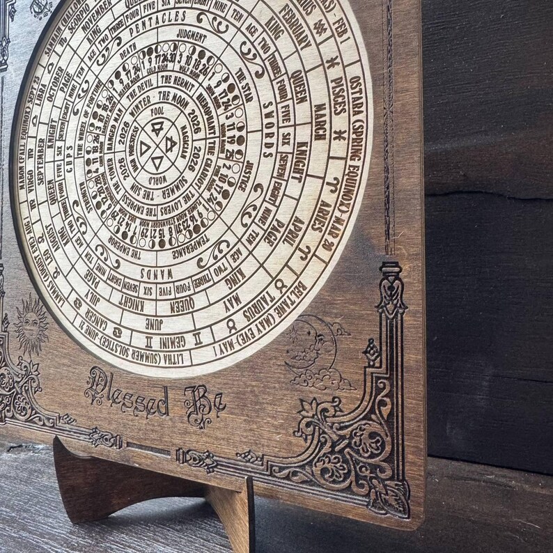 Może przedstawiać: Drewniany, okrągły kalendarz z symbolami astrologicznymi i tarota, w tym znakami zodiaku i nazwami kart. Kalendarz jest grawerowany ze skomplikowanymi detalami i słowami "Blessed Be". Jest wyświetlany na drewnianym stojaku.
