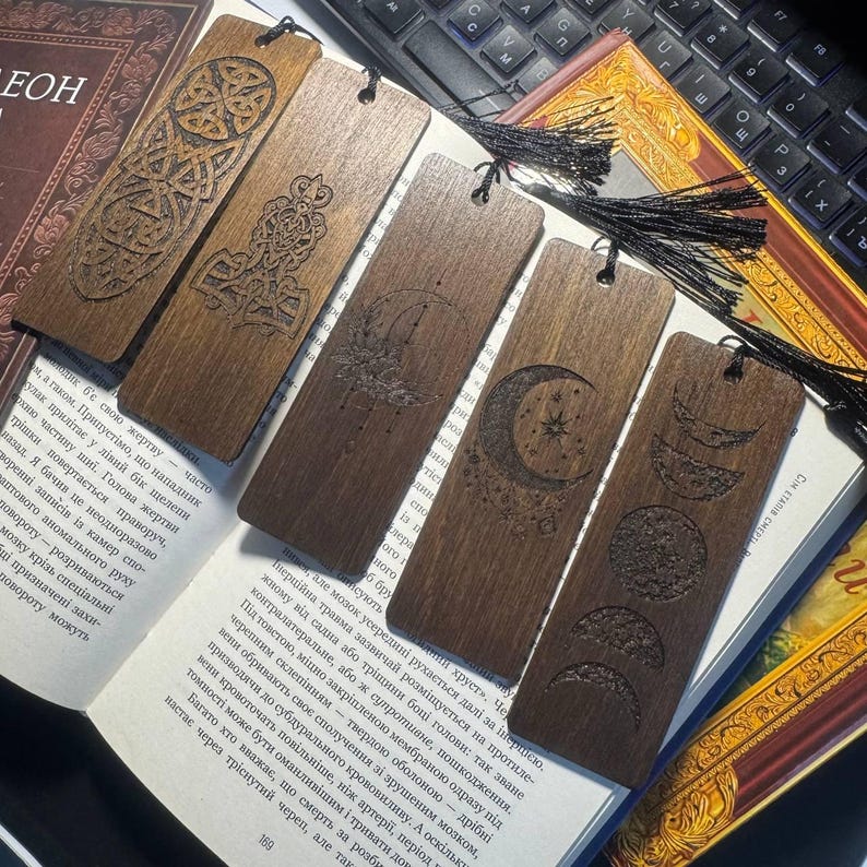 Pu&ograve; includere: Una collezione di segnalibri in legno con intricati disegni intagliati. I segnalibri presentano motivi celesti e a nodi celtici, con una finitura marrone scuro. Ogni segnalibro ha un nappa nera attaccata. Sono esposti su un libro aperto.
