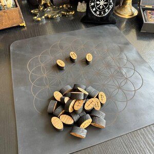May include: A collection of small, oval-shaped wooden tokens with black-stained sides and natural wood faces, each engraved with a unique rune symbol. Three tokens are placed separately above a larger pile of similar tokens on a black mat.