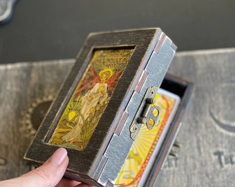 May include: A rectangular dark brown wooden box with a distressed finish and a metal latch. The lid features an inset illustration of an angel with red wings and the text "NOUVEAU TAROT". Inside the box is a deck of tarot cards with yellow designs.
