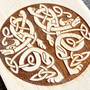 Può includere: Una placca di legno circolare con un design di nodo celtico dettagliato che presenta due animali intrecciati. Gli animali sono stilizzati e sembrano essere lupi o cani. Il design è intagliato nel legno ed è di colore marrone chiaro su uno sfondo marrone scuro.