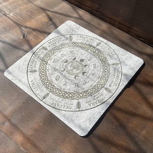 May include: A square light grey coaster featuring a circular design with the Wheel of the Year, including Samhain, Yule, Imbolc, Ostara, Beltane, Litha, Lughnasadh, and Mabon, alongside zodiac symbols, moon phases, and a central tree illustration.