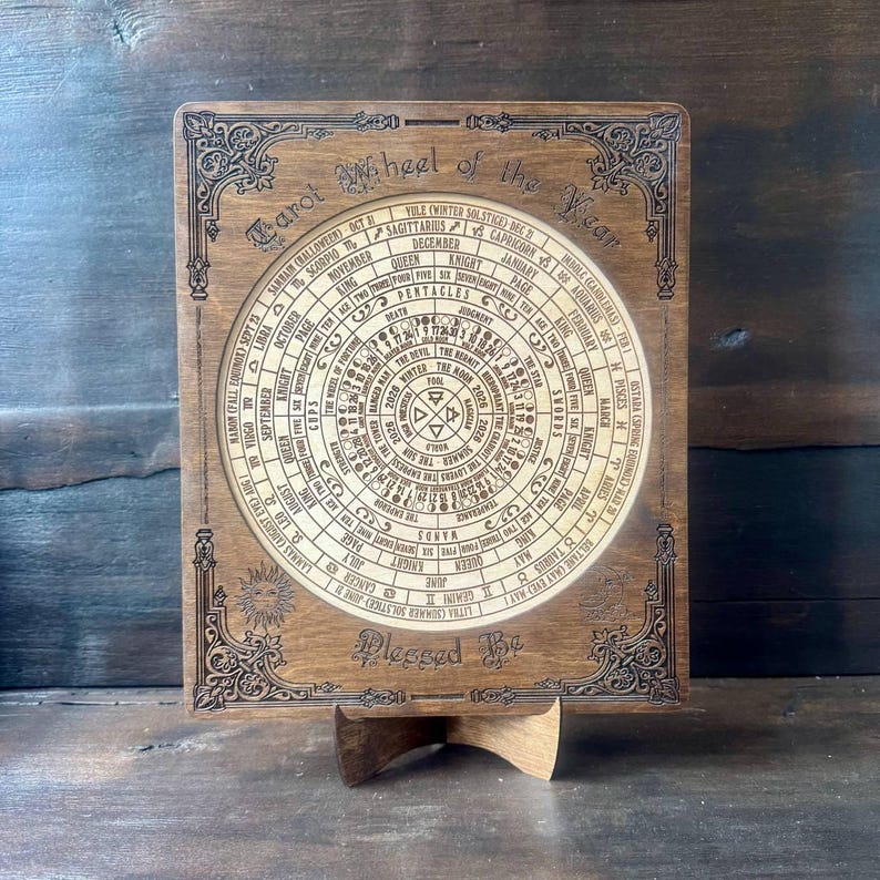 Może przedstawiać: Drewniane Koło Tarota Roku ze skomplikowanymi grawerowanymi detalami. Okrągły wz&oacute;r zawiera symbole astrologiczne, miesiące i nazwy kart tarota. Tekst "Blessed Be" znajduje się na dole. Drewniany element jest prezentowany na drewnianym stojaku.