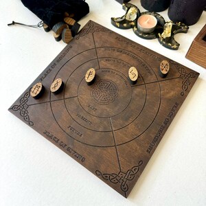 K&ouml;nnte beinhalten: Ein dunkelbraunes Holz-Divinationsbrett mit einge&auml;tzten keltischen Knotenmustern und kreisf&ouml;rmigen Zonen. Holz-Runensteine liegen auf dem Brett, im Hintergrund ein schwarzer Samtbeutel.