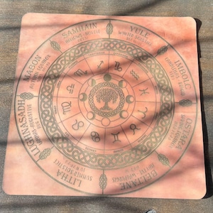 May include: A square, peach-colored mat featuring a circular design with the Wheel of the Year, zodiac signs, moon phases, and a central tree illustration. The text includes Samhain, Yule, Imbolc, Ostara, Beltane, Litha, Lughnasadh, and Mabon.