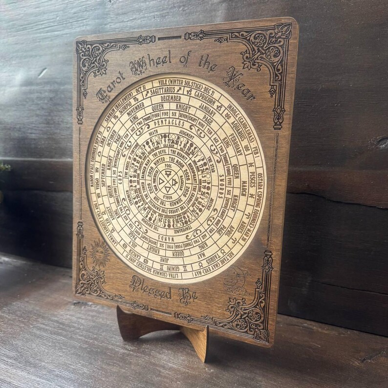 Może przedstawiać: Drewniane Koło Tarota Roku, z okrągłym wzorem z symbolami astrologicznymi i tekstem. Ciemnobrązowe drewno ma skomplikowane grawerowane detale i jest wyświetlane na drewnianym stojaku. Tekst "Blessed Be" znajduje się na dole.