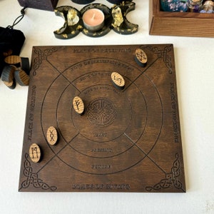 K&ouml;nnte beinhalten: Ein dunkelbraunes Holz-Divinationsbrett mit einge&auml;tztem keltischen Knoten und Runensymbolen. Das Brett hat konzentrische Kreise und Text, der Orte und Phasen angibt. Mehrere Runensteine aus Holz liegen auf dem Brett.