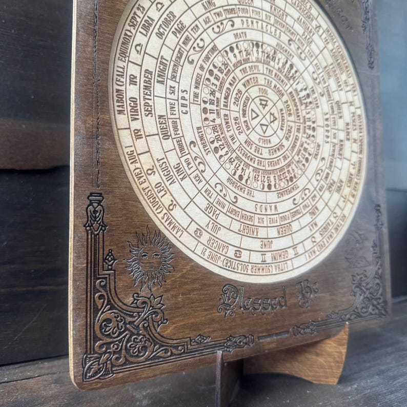 Może przedstawiać: Drewniany kalendarz dekoracyjny z okrągłym wzorem z symbolami astrologicznymi i tekstem. Kalendarz zawiera tekst "Blessed Be" i jest podparty drewnianym stojakiem. Wz&oacute;r jest wytrawiony w drewnie.