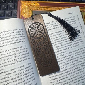 Pu&ograve; includere: Un segnalibro in legno scuro con un disegno a nodo celtico, appoggiato su un libro aperto. Il segnalibro ha un nappa nera. L'intricato lavoro a nodi &egrave; intagliato nel legno.