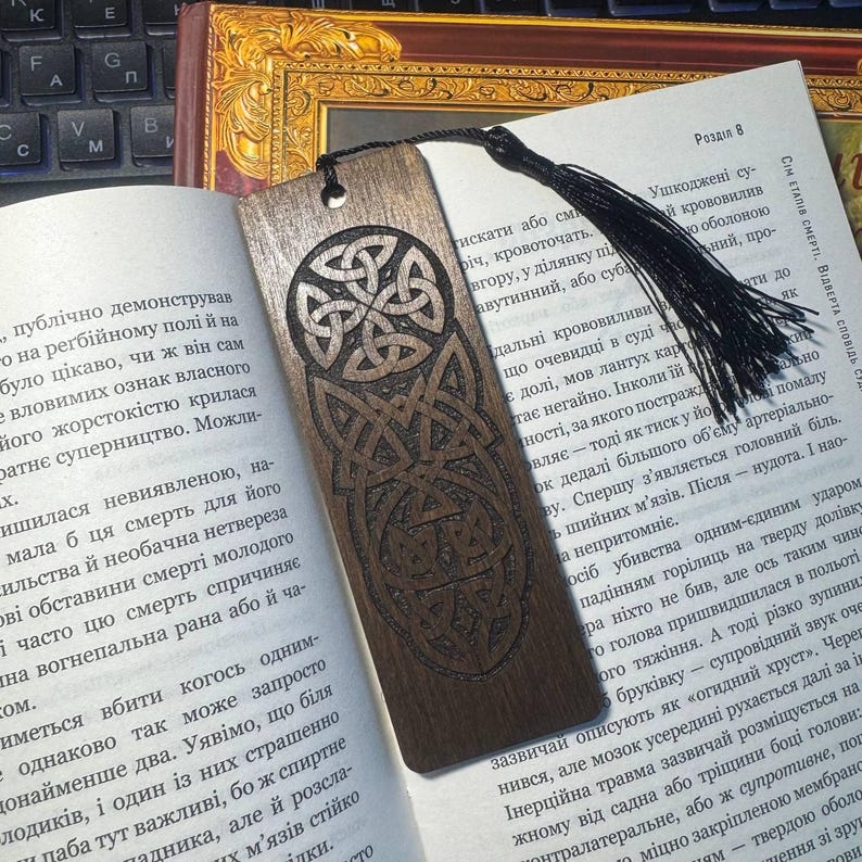 Pu&ograve; includere: Un segnalibro in legno scuro con un disegno a nodo celtico, appoggiato su un libro aperto. Il segnalibro ha un nappa nera. L'intricato lavoro a nodi &egrave; intagliato nel legno.