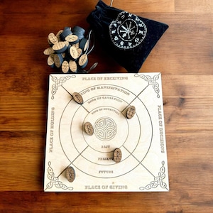 Könnte beinhalten: Ein hölzernes Divinationsbrett mit eingeätzten keltischen Knotenmustern und Text, darunter "Place of Receiving" und "Place of Giving". Enthält Runensteine aus Holz und einen schwarzen Samtbeutel mit weißem astrologischem Design.